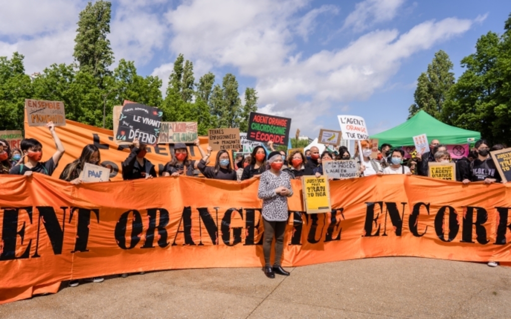 Photo d'une mobilisation - Tran to Nga devant une banderole : "L'agent orange tue encore" et derrière des militant.e.s avec des pancartes